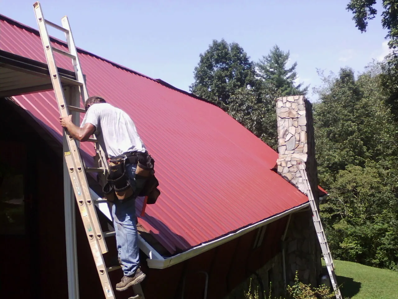 Roofer on a ladder beside a red metal roof and stone chimney — Soffit & Fascia Repair by Roofing Brothers in Elkhorn City